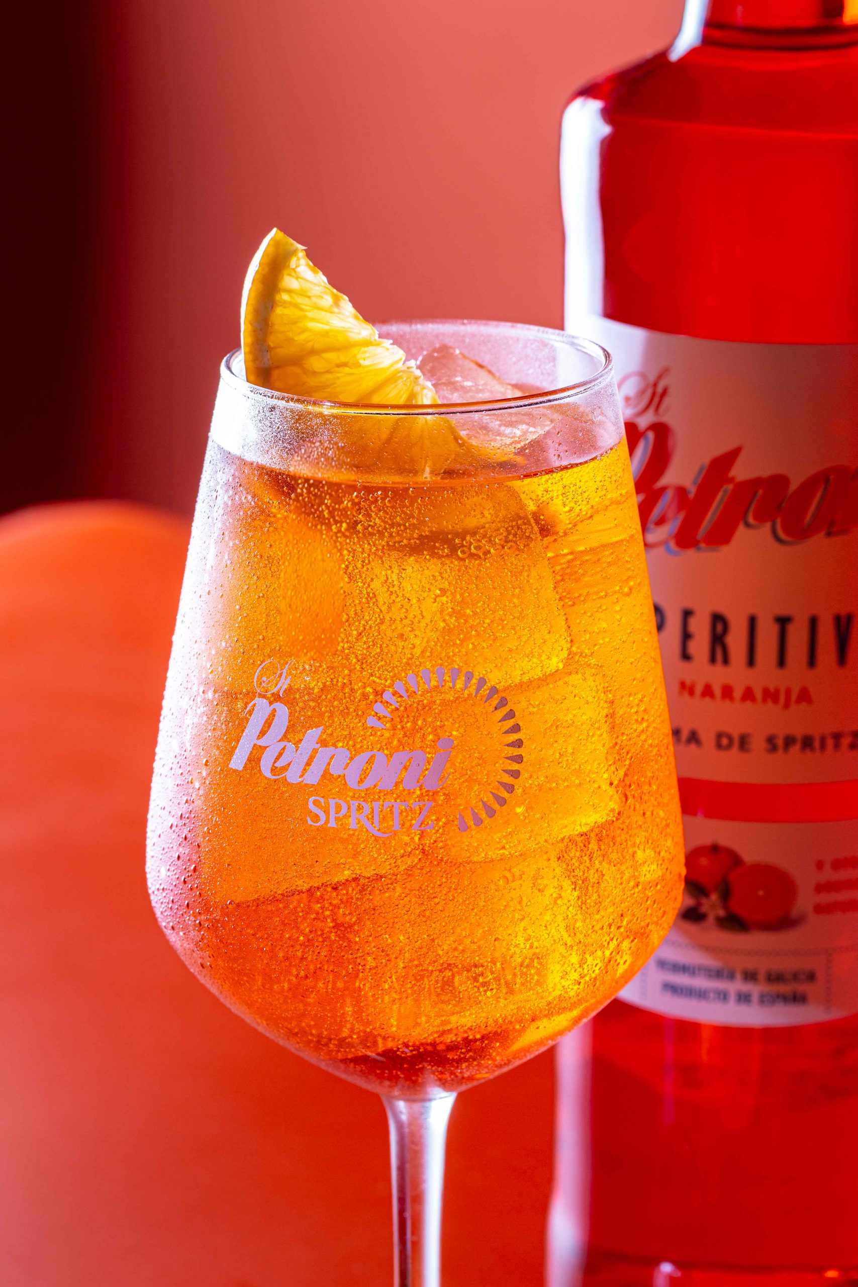 Copa de Petroni Spritz con hielo y rodaja de naranja junto a la botella del aperitivo, sobre fondo rojo brillante